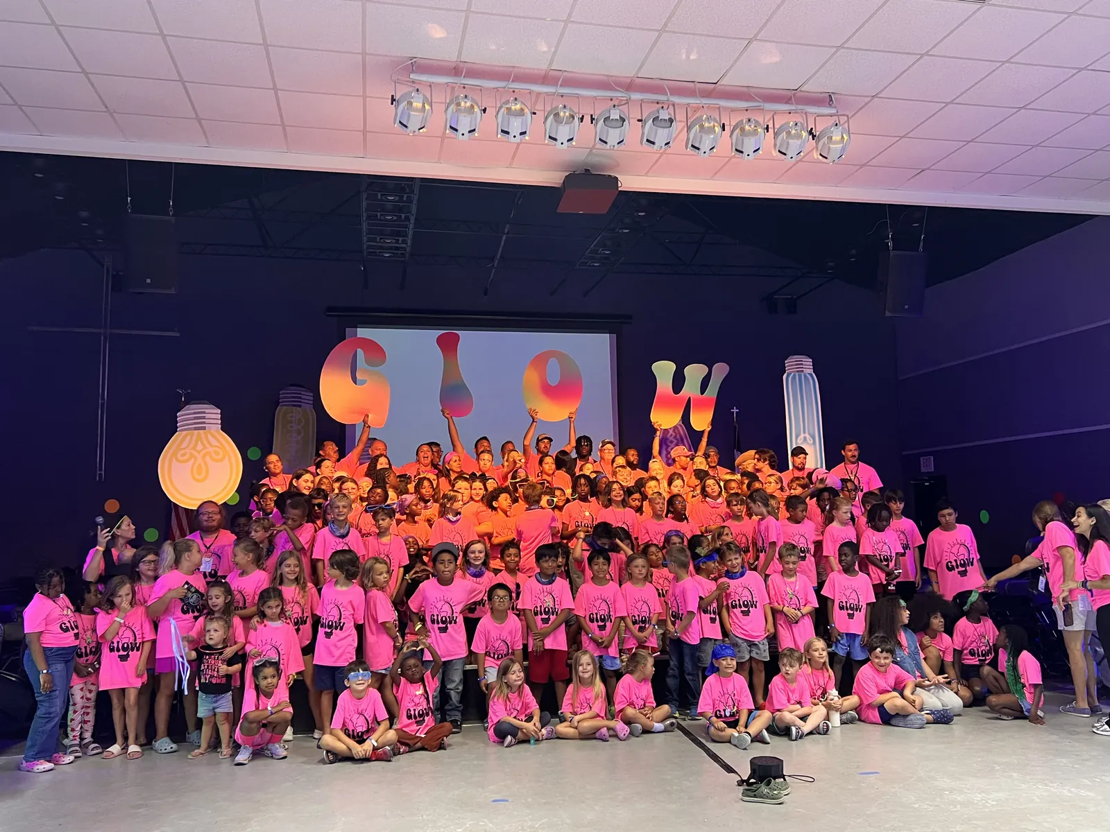 The whole camp in pink GLOW shirts posing in front of the GLOW stage